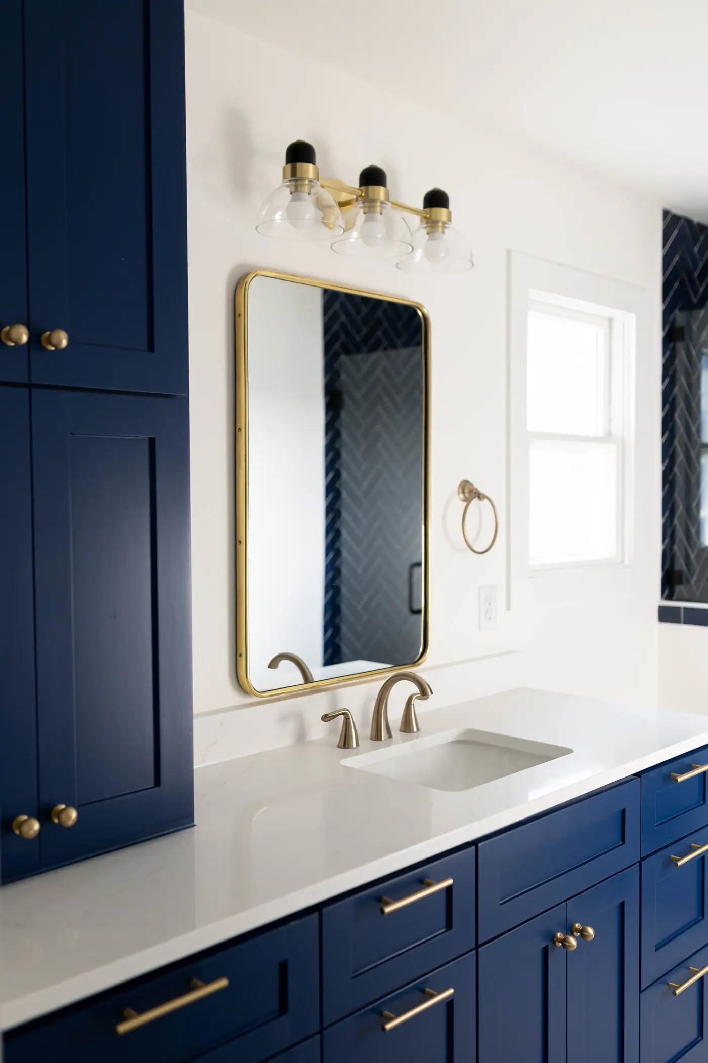 A navy tall cabinet with brass knobs, a brass-framed mirror reflecting the navy herringbone shower, and a champagne bronze faucet, in a 6th Avenue Homes bath remodel