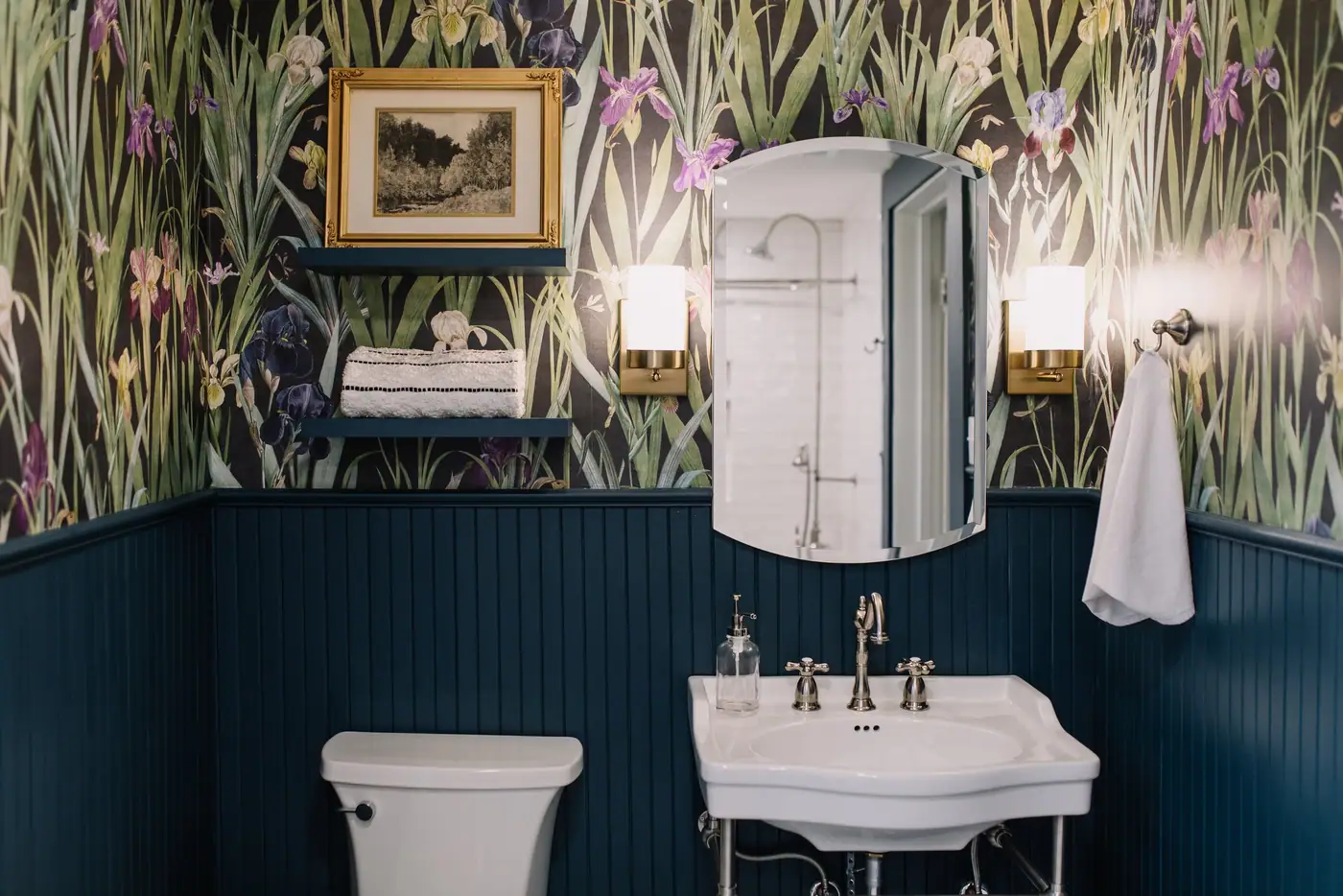 A small bath with floral wallpaper over navy beadboard, a console sink, and a beveled mirror between two brass sconces in Fairmount