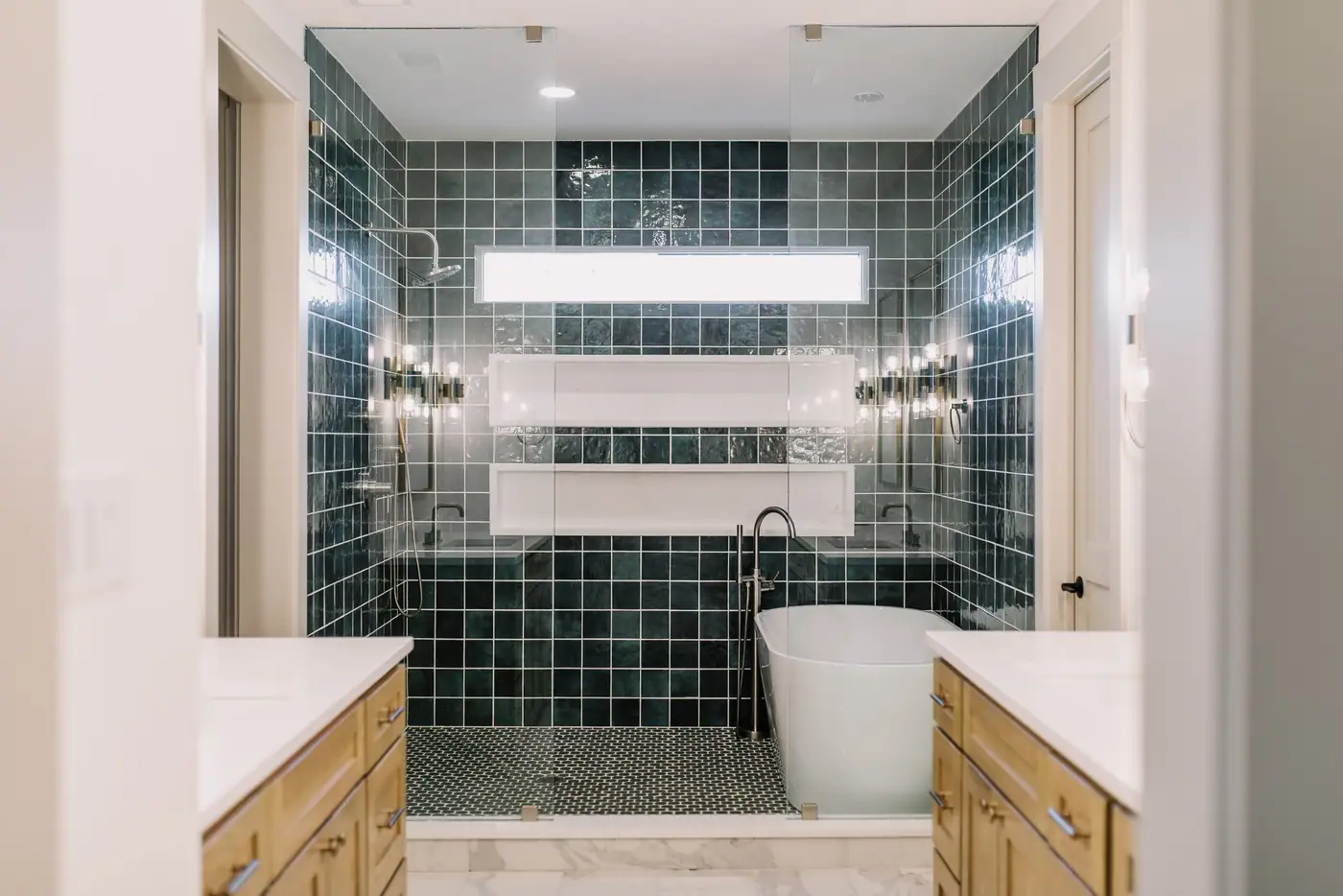 A wet room in dark green square tile with a freestanding tub and a rain shower between twin wood vanities in a 6th Avenue Homes bath