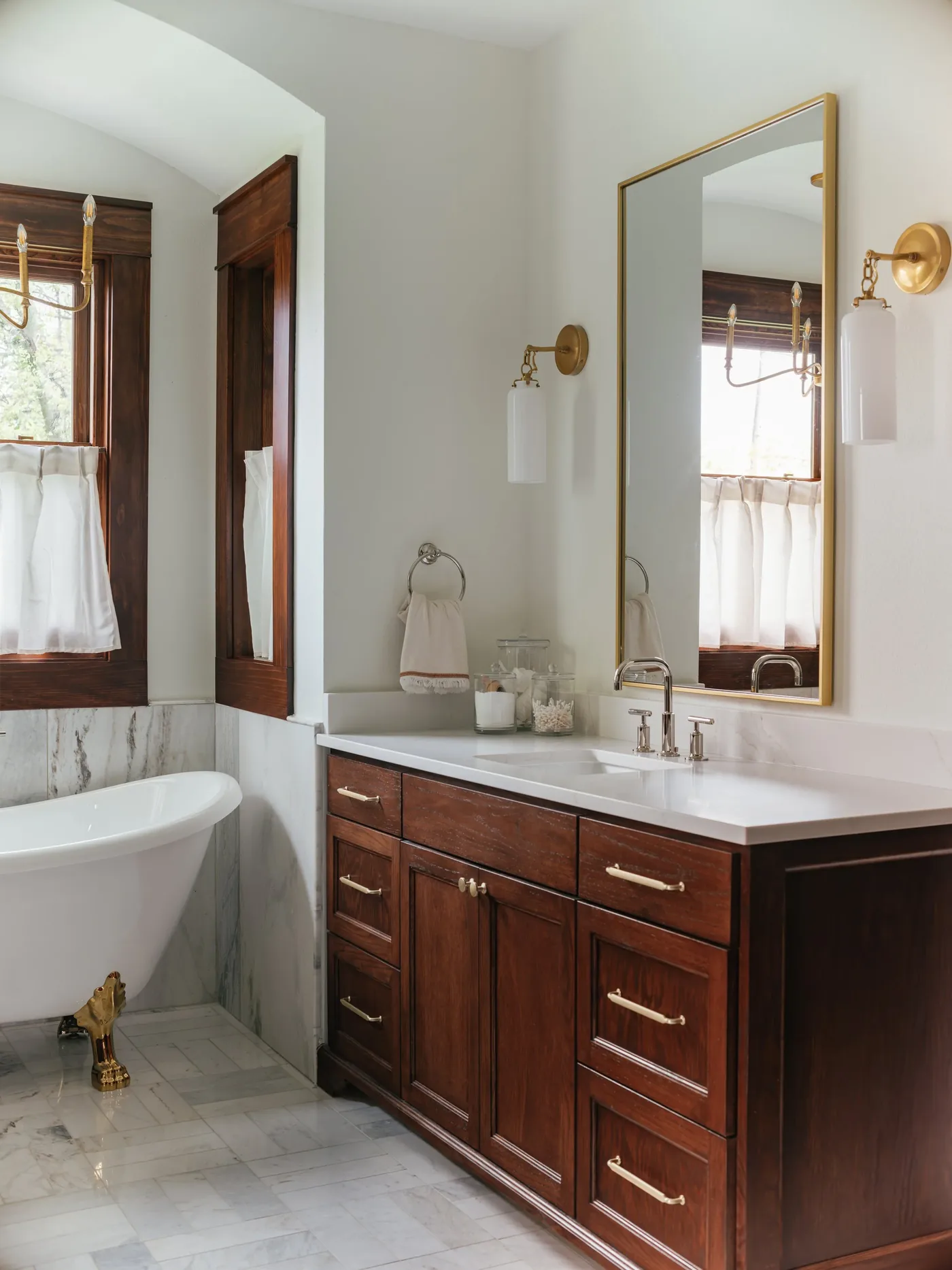 A stained wood vanity with brass pulls under a brass-framed mirror beside a clawfoot tub in a Ryan Place primary bath