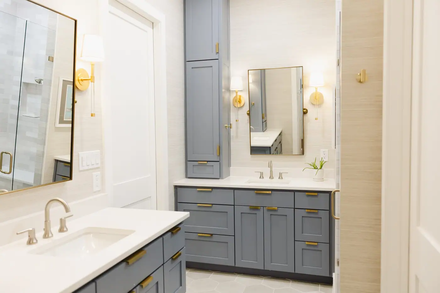 A smoky blue vanity and tall cabinet with brass pulls, a brass-framed mirror, and two sconces on a textured wall in a Tanglewood bath