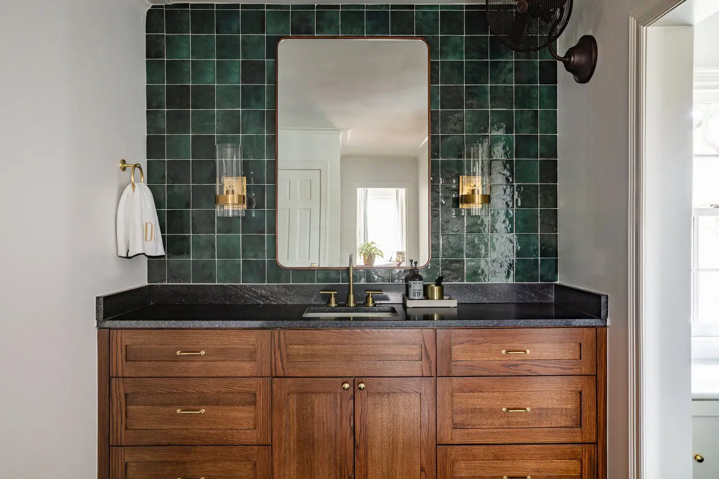 A stained wood vanity with a black counter and brass pulls under a slim bronze-toned mirror between two glass cylinder sconces, against a green square tile wall, in a 6th Avenue Homes primary suite bath